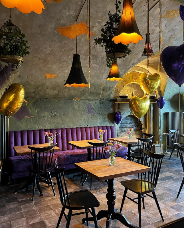 Na obrazie widzimy przytulne wnętrze restauracji, które ma rustykalny, a zarazem elegancki charakter. W centralnym punkcie znajduje się fioletowa kanapa, otoczona drewnianymi stolikami i czarnymi krzesłami. Na każdym stoliku stoją wazony z kwiatami, dodając wnętrzu uroku.