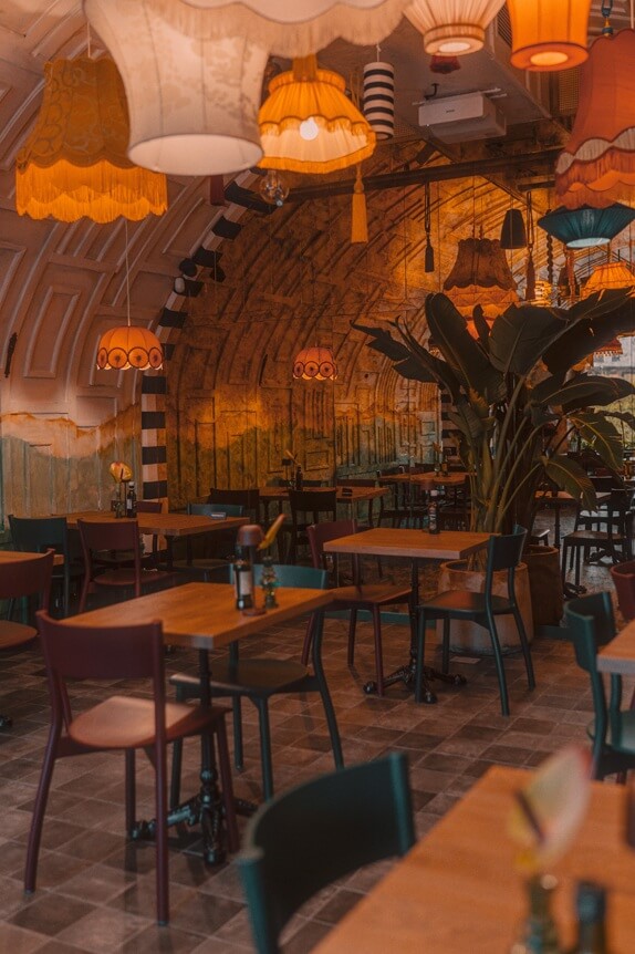 Na obrazku widać wnętrze przytulnej restauracji. Dominują drewniane stoły i krzesła starannie ustawione, a na niektórych stołach znajdują się przyprawy i drobne dekoracje. Z sufitu zwisają kolorowe i oryginalne abażury, które nadają przestrzeni ciepłą i klimatyczną atmosferę. Ściany są artystycznie wykończone, z mieszanką malowanych i teksturowanych powierzchni — na przykład charakterystyczna kolumna w paski. Całości dopełnia duża roślina, która wnosi nieco zieleni i świeżości do wnętrza.
