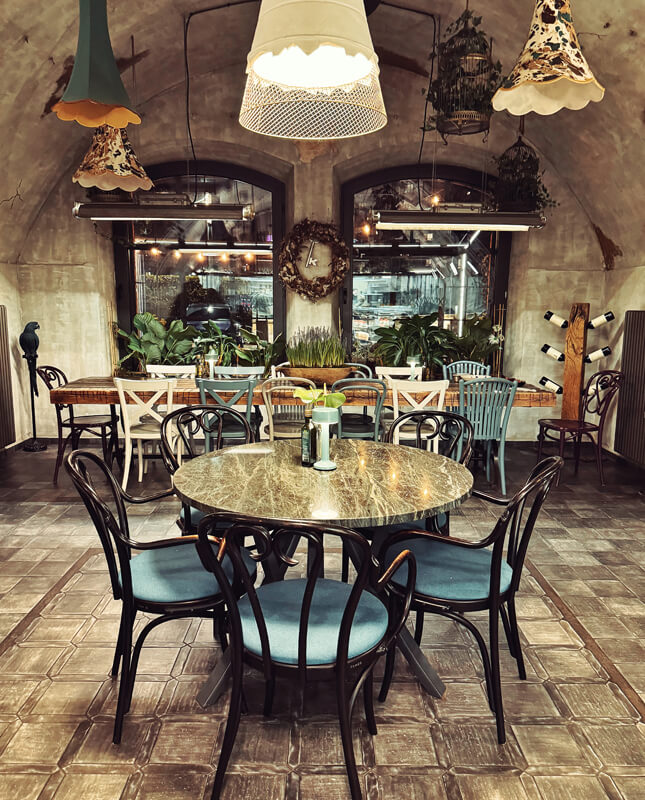 Zdjęcie przedstawia klimatyczne wnętrze restauracji o przytulnym, rustykalnym charakterze. W centralnej części kadru znajduje się okrągły stół otoczony klasycznymi krzesłami z ciemnego drewna i tapicerką w odcieniu niebieskim. W tle widoczny jest długi stół ustawiony przy dużych oknach z łukowatymi nadprożami, za którymi rozpościera się widok na kuchnię lub ogród zimowy. Otoczenie obfituje w rośliny doniczkowe i subtelne dekoracje – wianki, drewniane akcenty i designerskie lampy wiszące w różnych stylach.