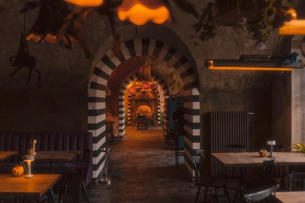 Obrazek przedstawia wnętrze restauracji o wyjątkowym i intrygującym designie. Głównym elementem są łukowate przejścia w czarno-białe pasy, które tworzą efekt tunelu i przyciągają wzrok w głąb przestrzeni. Oświetlenie jest ciepłe i przytłumione, z pomarańczowymi odcieniami pochodzącymi z wiszących lamp, co dodaje przytulnej i tajemniczej atmosfery. Na pierwszym planie znajdują się stoły i krzesła, a na stolikach leżą małe dynie, co sugeruje sezonowy motyw.