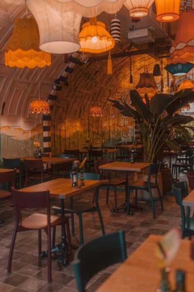 Na zdjęciu przedstawiono wnętrze nastrojowej, kameralnej restauracji. Przestrzeń zdominowana jest przez drewniane stoły i krzesła, starannie rozstawione, z dodatkami w postaci przypraw i subtelnych dekoracji. Z sufitu zwisają nietuzinkowe, kolorowe abażury, których ciepłe światło buduje przyjemny, klimatyczny nastrój. Ściany ozdobione są w artystyczny sposób — łączą malowane fragmenty z fakturami, w tym wyróżniającą się kolumną w pasy. Całość aranżacji dopełnia duża roślina, która ożywia wnętrze i wprowadza naturalny akcent.