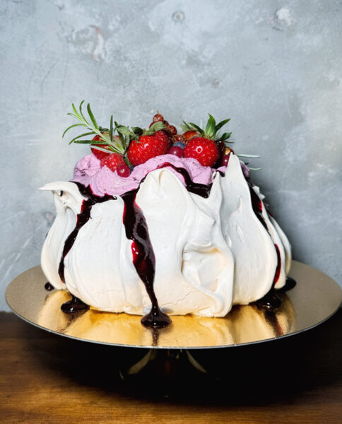 Zdjęcie przedstawia okrągły tort bezowy w kształcie gniazda, umieszczony na złotej, metalowej podstawce. Beza ma nieregularny, pofalowany kształt i jest hojnie polana ciemnym sosem, który spływa po jej bokach. Wewnątrz "gniazda" znajduje się krem w fioletowo-różowym kolorze, a na nim ułożone są świeże truskawki, borówki oraz gałązki rozmarynu. Tort stoi na drewnianym blacie, na tle szarej, teksturowanej ściany.