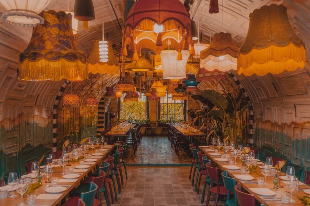 Na zdjęciu widzimy wnętrze restauracji o wyjątkowym, artystycznym charakterze. Sufit zdobią różnorodne, vintage’owe abażury w różnych kształtach i kolorach, które tworzą ciepłą, przytulną atmosferę. Ściany mają rustykalną fakturę, uzupełnioną malowidłami i dekoracyjnymi panelami, co dodaje przestrzeni kreatywnego wyrazu. Stoły są długie, starannie nakryte talerzami, szkłem i sztućcami — wszystko wskazuje na przygotowanie do większego spotkania.