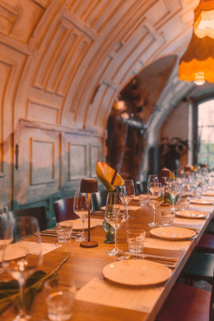 Na zdjęciu widzimy elegancko nakryty, długi stół w restauracji lub sali bankietowej, przygotowany na uroczystą kolację lub przyjęcie. Na stole znajdują się starannie ułożone talerze, kieliszki do wina i wody, sztućce oraz drobne dekoracje — kwiaty i świece — które dodają całości wyrafinowanego charakteru. Wnętrze zdobi sklepiony sufit z ozdobnymi panelami, a ciepłe, nastrojowe oświetlenie tworzy przytulną, świąteczną atmosferę. To przestrzeń, która łączy klasyczną elegancję z gościnnym klimatem, idealna na wyjątkowe spotkania.