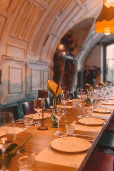 Na zdjęciu widzimy elegancko nakryty, długi stół w stylowym wnętrzu restauracyjnym lub sali bankietowej. Każde miejsce wyposażone jest w starannie ułożone talerze, kieliszki do wina i wody, sztućce oraz dekoracyjne elementy — wśród nich małe wazony z kwiatami i świeczniki, które tworzą przytulny, wyrafinowany klimat. W tle widoczny jest sklepiony sufit o teksturowanej powierzchni, subtelnie oświetlony ciepłym światłem.