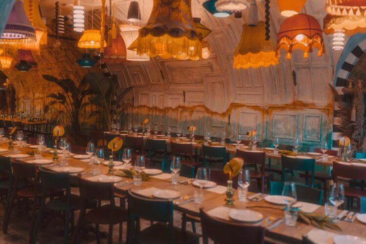 Na zdjęciu widoczna jest przestronna sala restauracyjna przygotowana na przyjęcie, z długimi stołami starannie nakrytymi zastawą i szkłem. Nad nimi wiszą dekoracyjne lampy o ciepłym świetle.